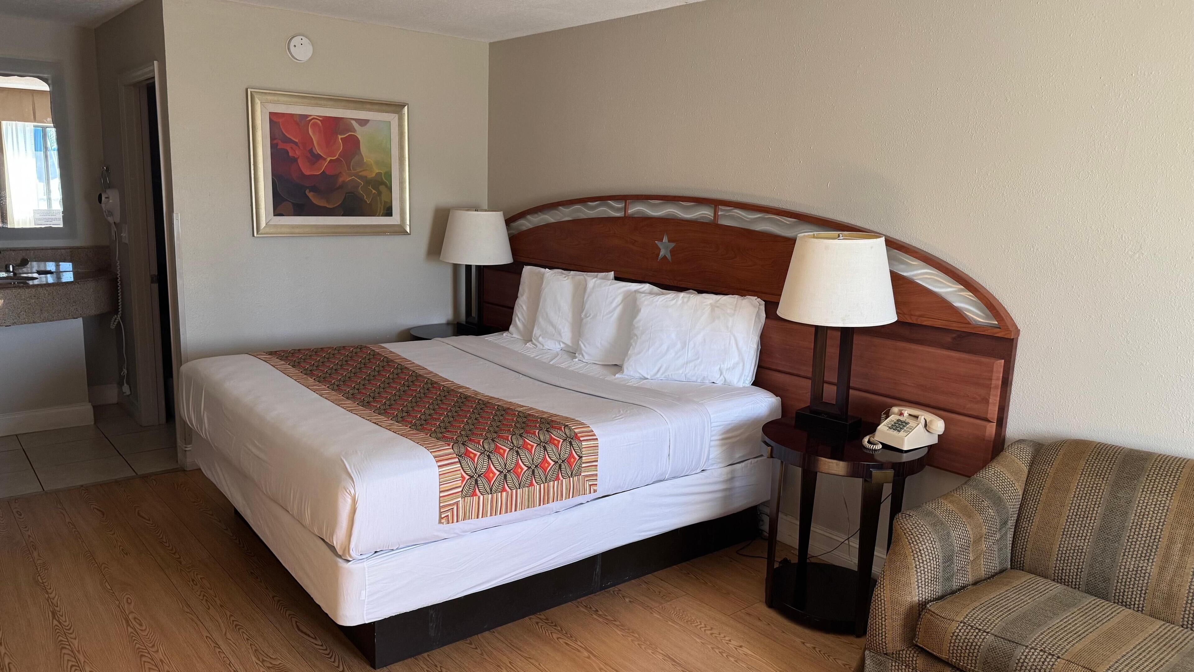 Habitación de hotel con cama, dos lámparas de noche, teléfono, silla y un cuadro en la pared.