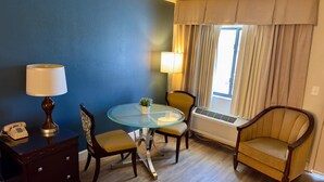 A hotel room with a blue wall, a glass table with chairs, a telephone, a lamp, a potted plant, and a window with curtains. - Hotel MonteCarlo (Kissimmee)