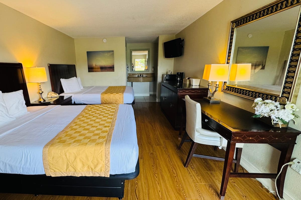 A hotel room with two beds, a desk, a chair, a mirror, and a small kitchenette.