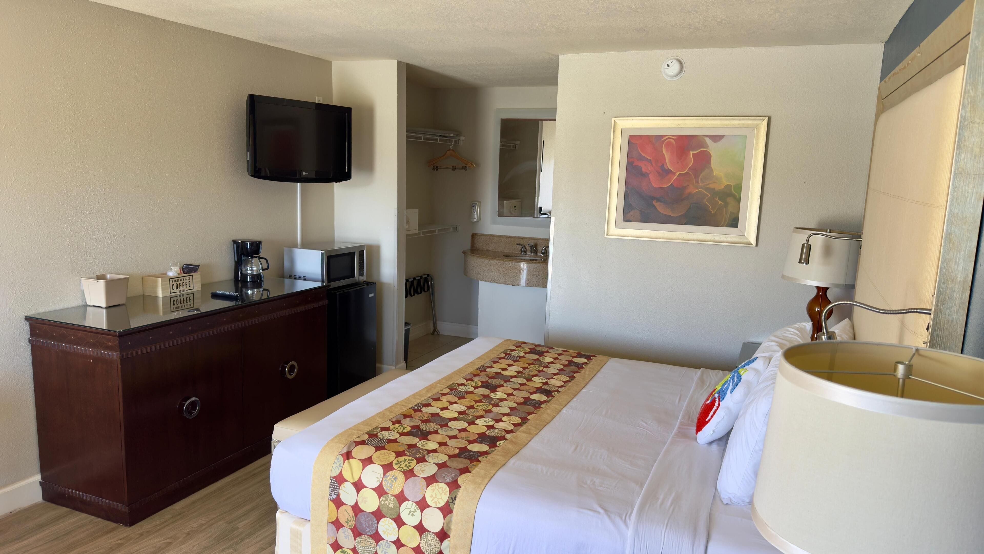 Quarto de hotel com cama, penteadeira, televisão, micro-ondas, cafeteira e um quadro na parede.