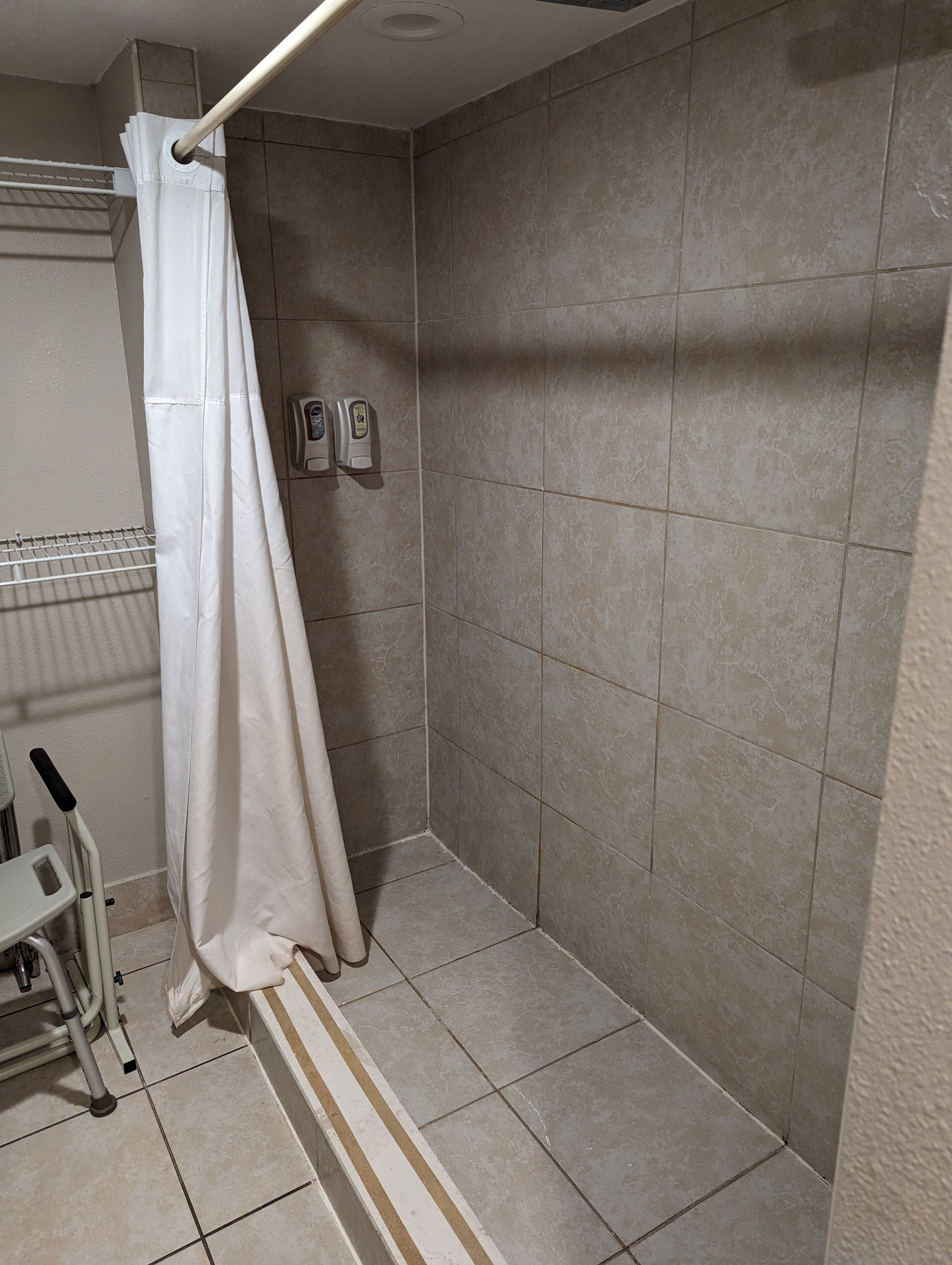 Un área de ducha con azulejos color beige, una cortina de ducha blanca y un dispensador de jabón en la pared.