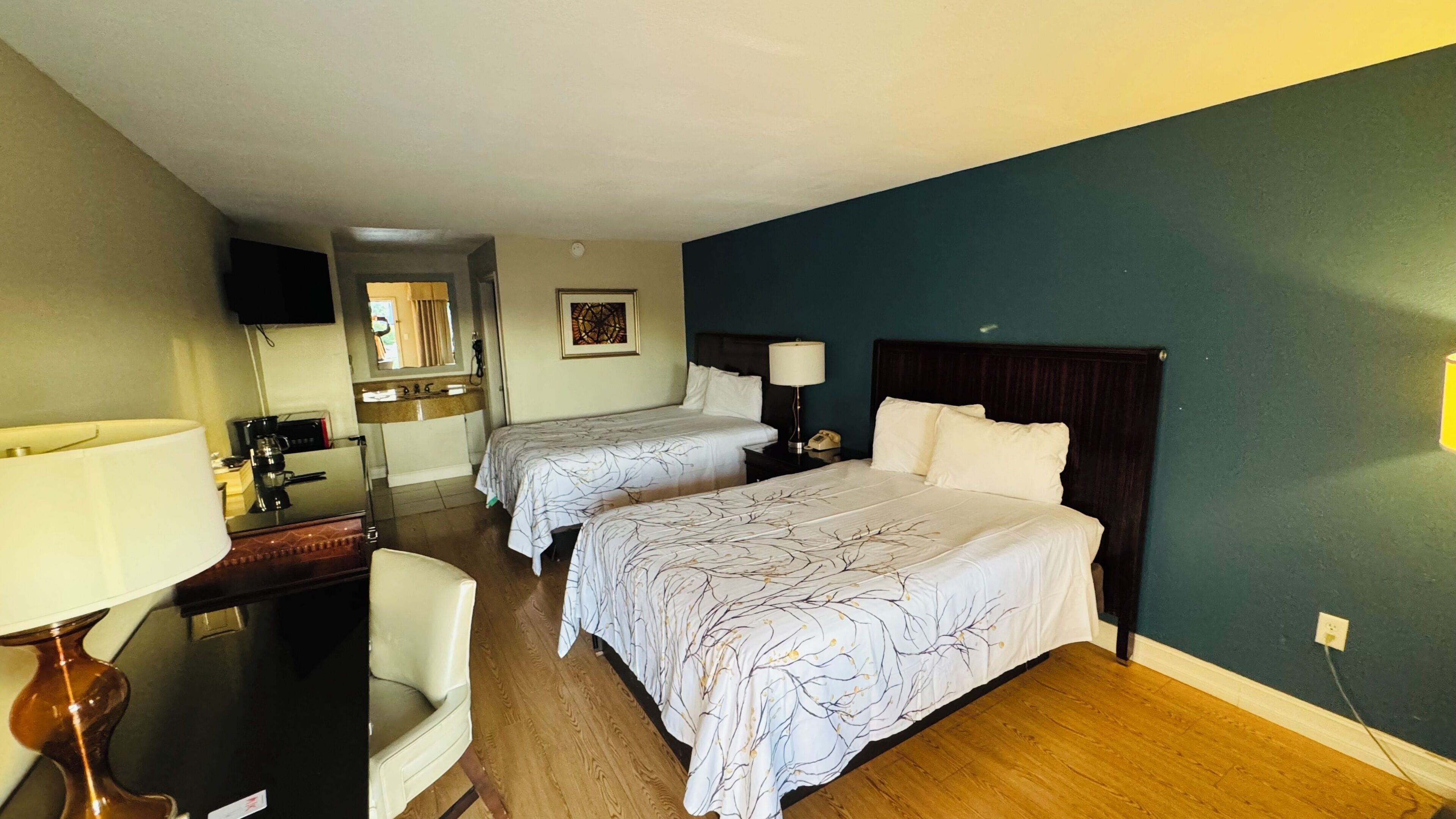Une chambre d’hôtel avec deux lits, un bureau, une chaise, une lampe de bureau, un téléphone et un tableau au mur.