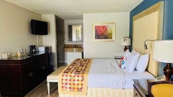 Habitación de hotel con cama, mesita de noche, lámpara, televisión y una zona de kitchenette.