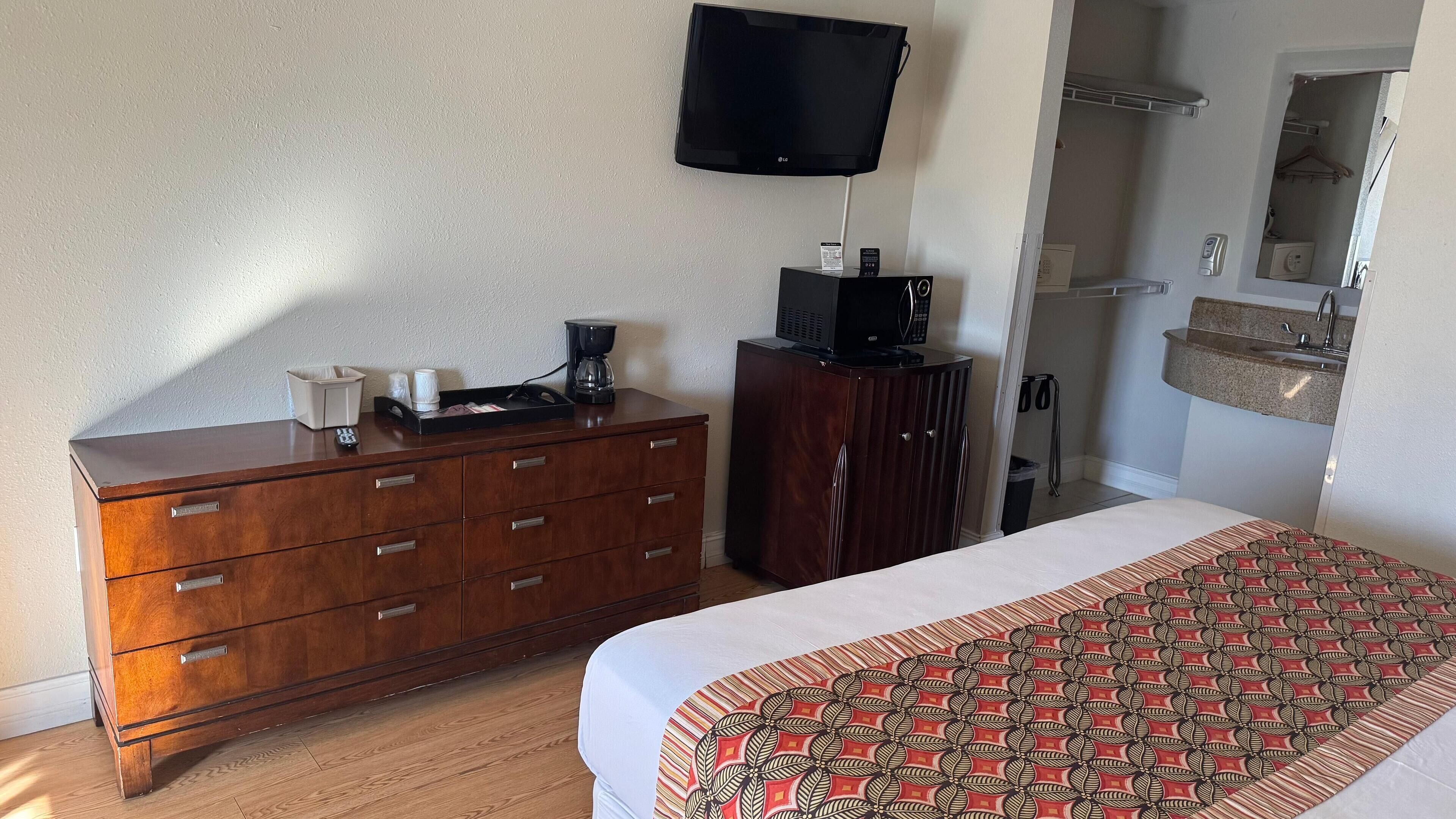 Een hotelkamer met een houten dressoir, een bed met een bedsprei met een patroon, een televisie aan de muur en een klein kitchenette.