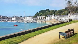 A coastal promenade with a bench, a stone wall, and a marina with boats.