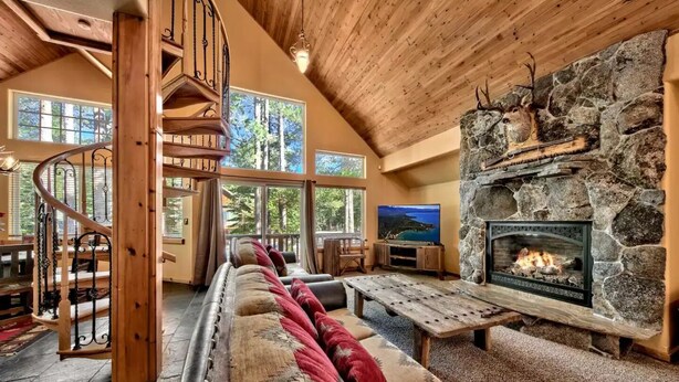 A spacious living room with a stone fireplace, a wooden staircase, and large windows offering a view of the outdoors. - Luxury House, Hot Tub, Pet Friendly, Pool Table, EV Charger
 (South Lake Tahoe)
