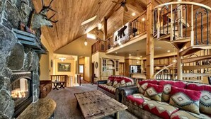 A rustic interior with a stone fireplace, a wooden ceiling, a large sofa, and a staircase with a balcony. - Luxury House, Hot Tub, Pet Friendly, Pool Table, EV Charger
 (South Lake Tahoe)