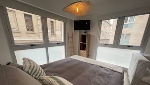 Ein Schlafzimmer mit einem großen Bett, einem Fernseher und Blick auf Gebäude durch die Fenster.