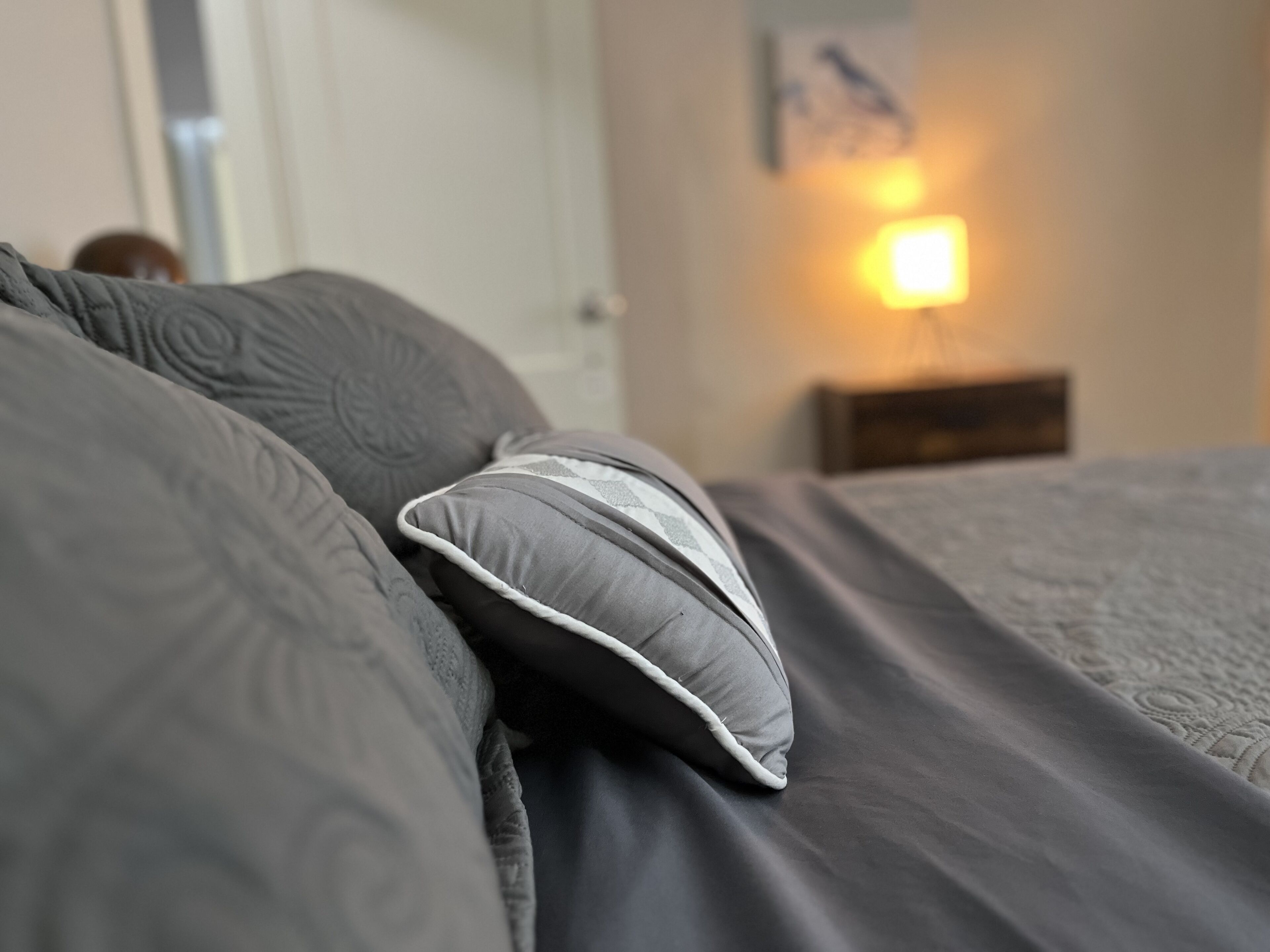 Un letto con biancheria e cuscini grigi, un comodino con una lampada e una porta sullo sfondo.