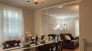 Ein Essbereich mit Holztisch, Stühlen und Glasvasen mit Blumen. Auf dem Tisch steht eine Weinflasche. Im Hintergrund sind ein Kronleuchter, eine Lampe und ein Sofa zu sehen.