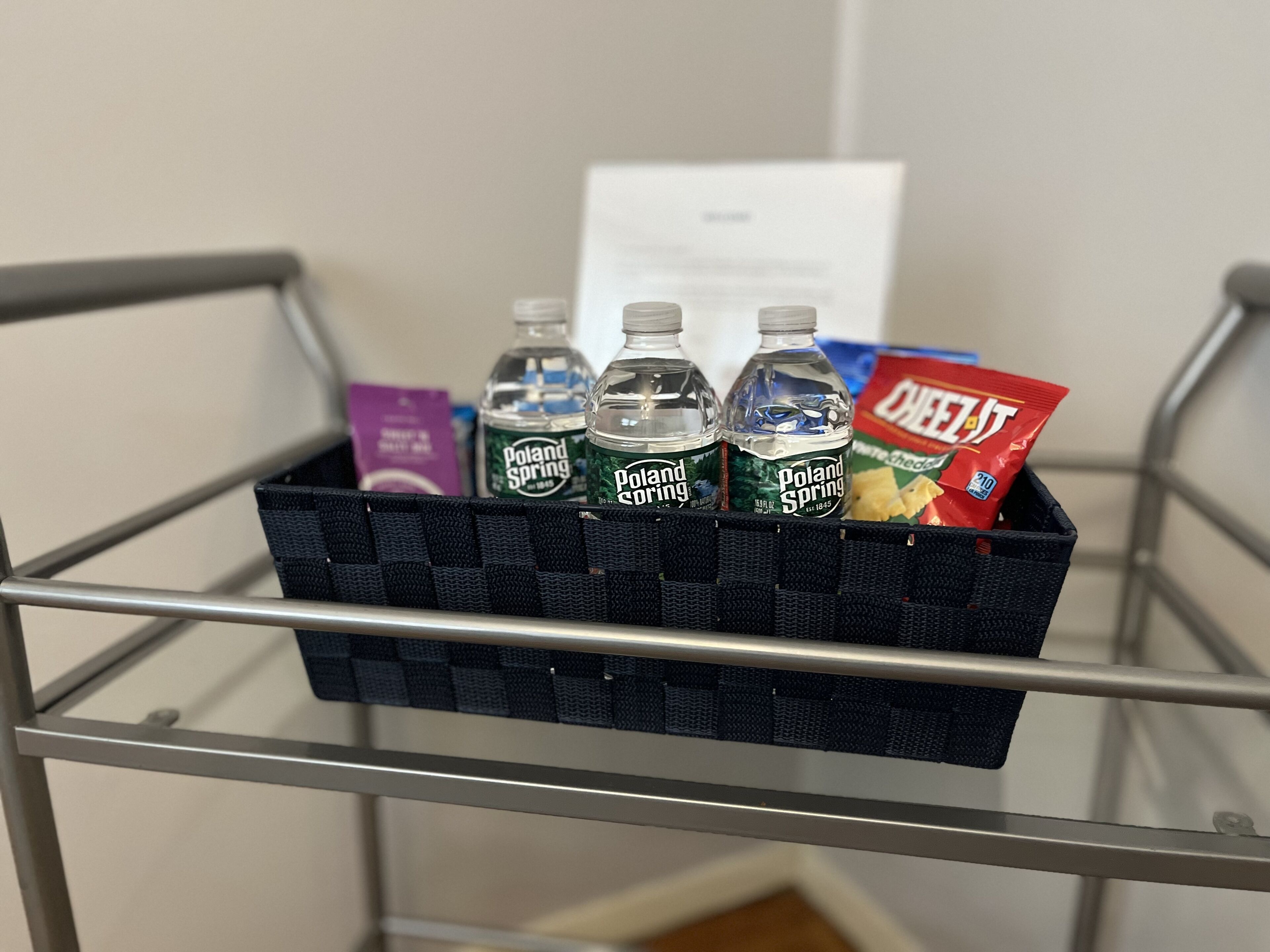 A metal shelf with a black basket containing bottles of Poland Spring water, a purple box, and a red bag of chips.