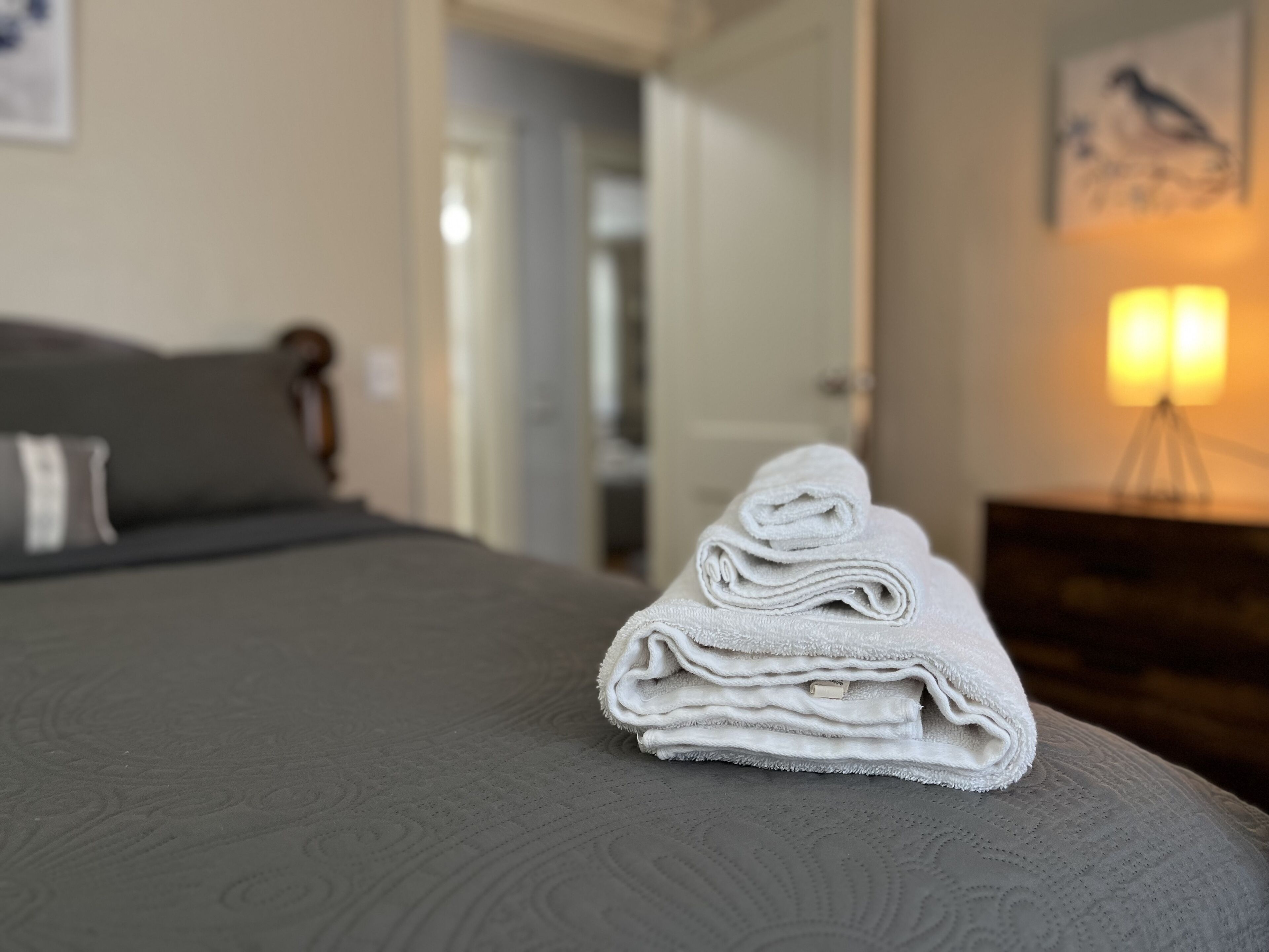Ein ordentlich bezogenes Bett mit einem gefalteten weißen Handtuch darauf, einem grauen Kopfteil und einer Wandlampe.