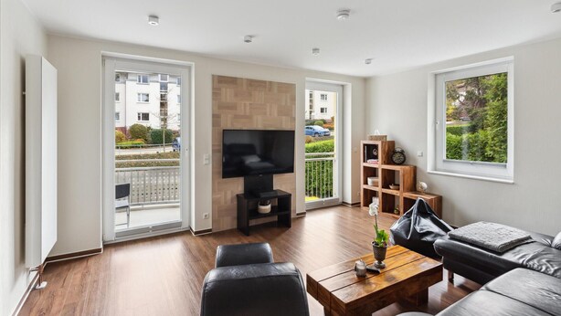 Un salón moderno con televisor de pantalla plana, sofá de cuero, mesa de centro de madera y un balcón con vistas a una zona residencial.