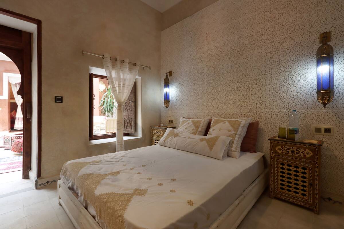 Un dormitorio con una cama grande, una mesita de noche, una lámpara de pared y un ventanal con cortina.