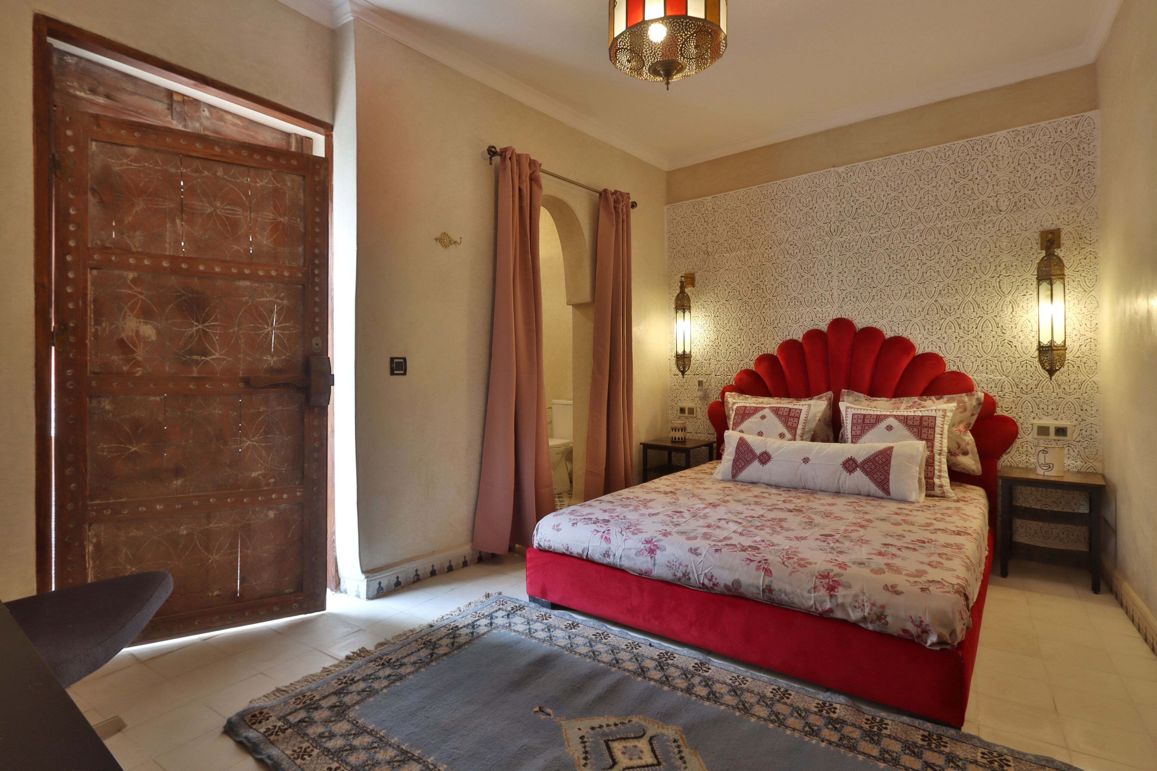 A bedroom with a large bed, a wooden door, a patterned rug, and two wall sconces.