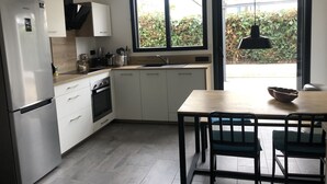 A modern kitchen with white cabinets, a stainless steel refrigerator, a dining table with chairs, and a large window with a view of greenery outside. - BIG BEN - à 750m de la Plage (Les Sables-d´Olonne)