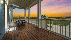 A wooden deck with a table and chairs, overlooking a residential area during sunset. - Modern Beach Home with Stunning West Bay Views (Galveston)