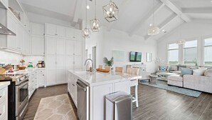 A modern kitchen with white cabinets, stainless steel appliances, and a central island. There is a dining area with a sofa and a TV. - Modern Beach Home with Stunning West Bay Views (Galveston)