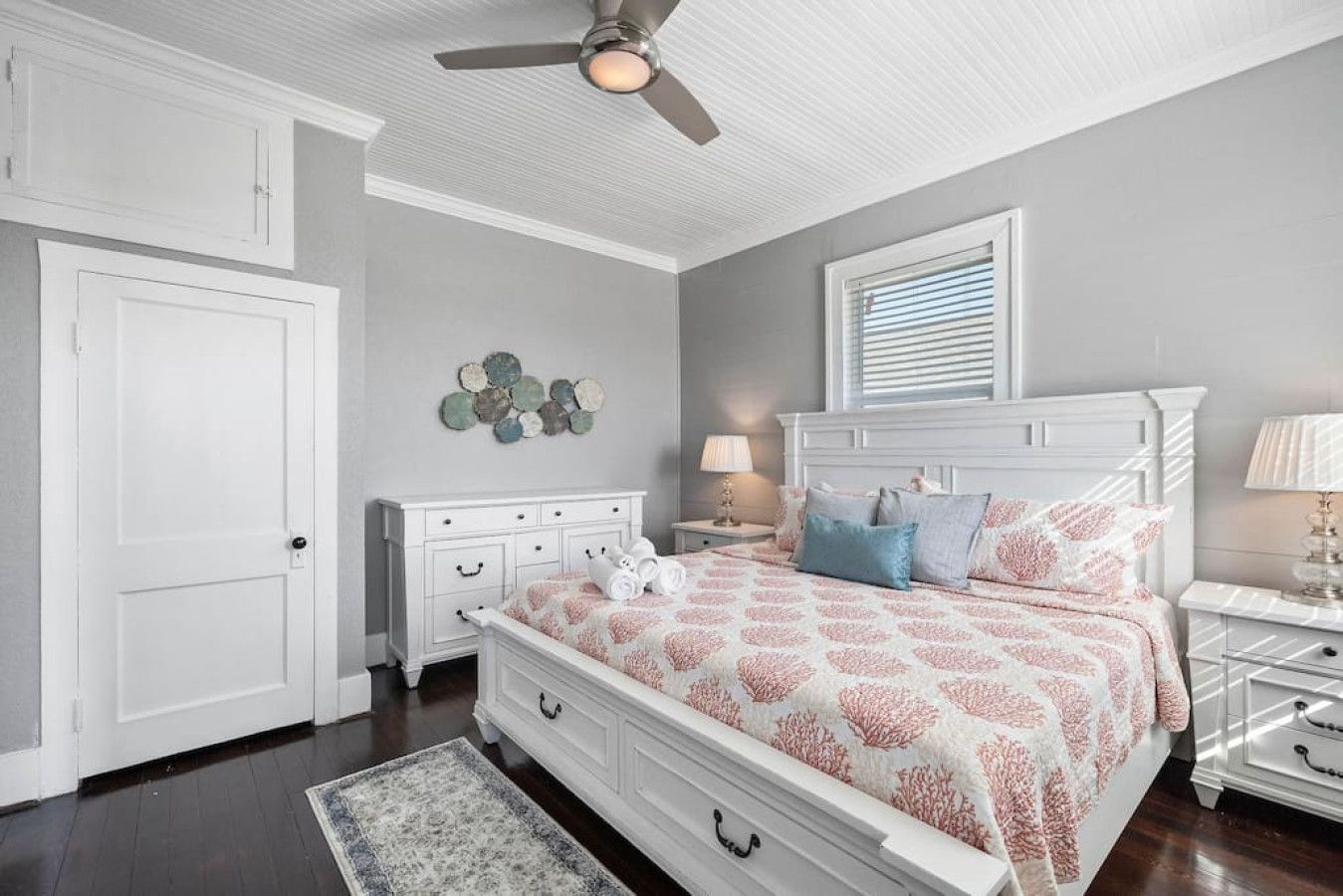 Une chambre à coucher comprenant un lit, des tables de chevet, une commode, un ventilateur de plafond et une fenêtre avec des stores.