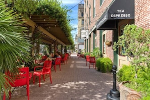 Un café en plein air avec des chaises et des tables rouges, un bâtiment en briques sur lequel est apposée une enseigne indiquant un salon de thé et un café proposant des expressos, et de la verdure tout autour.