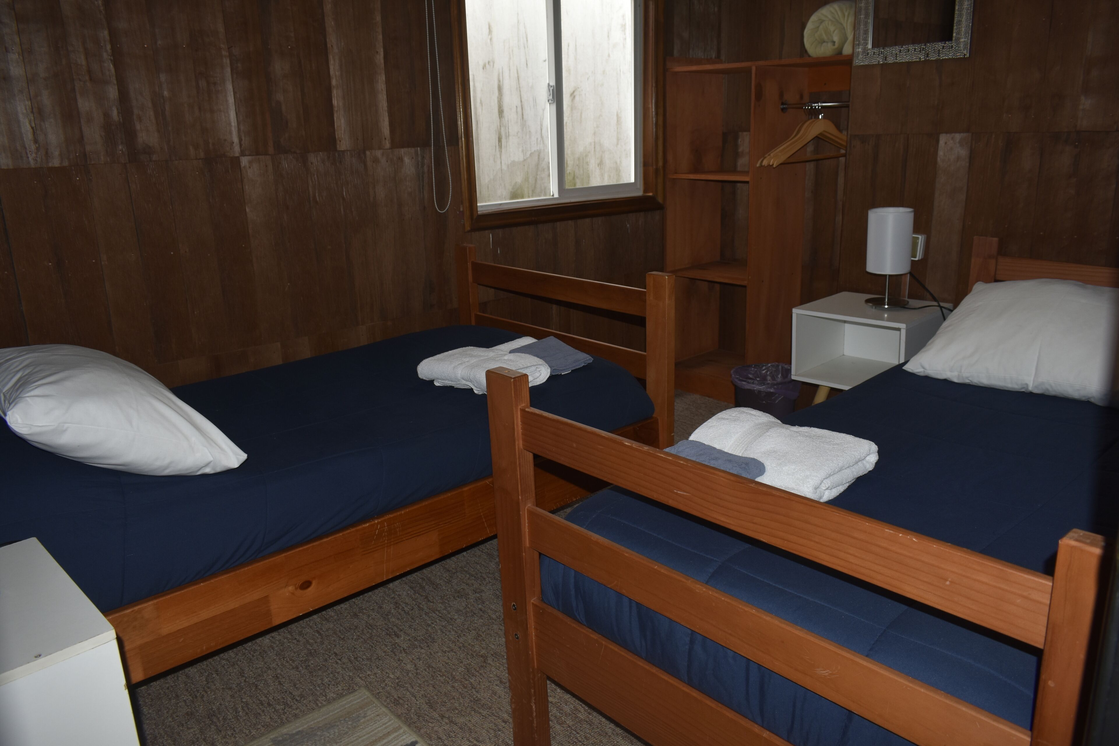 A room with two wooden bunk beds, a small bedside table with a lamp, and a window with a view of the outside.