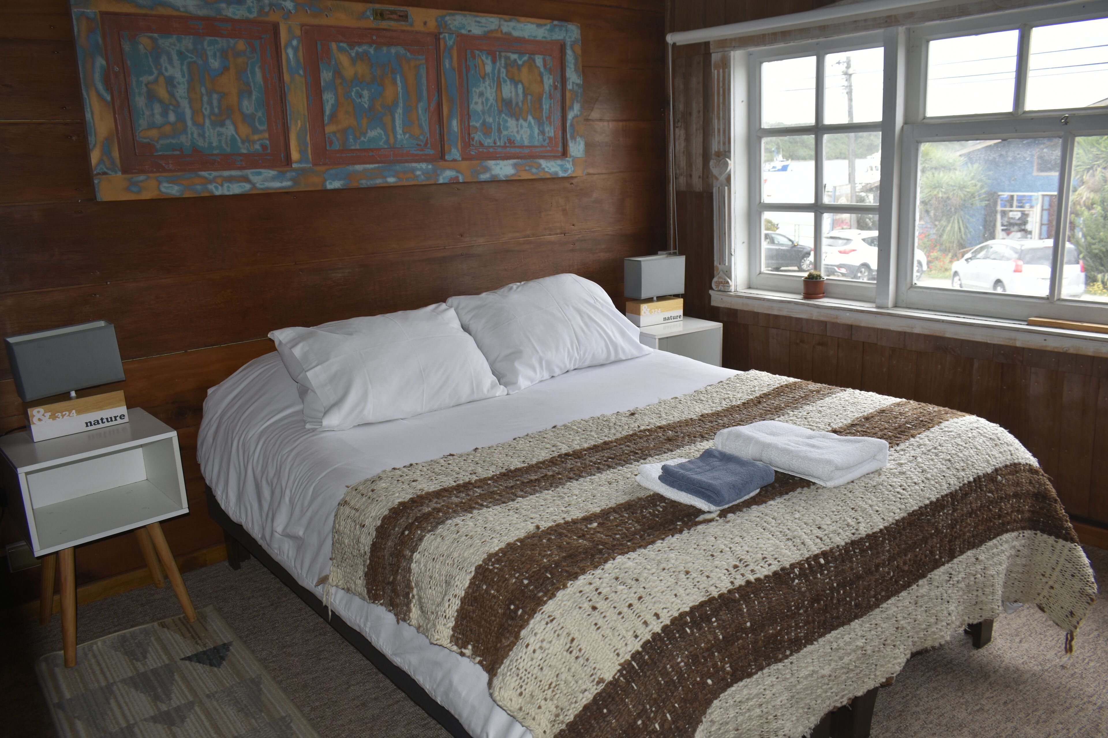 Une chambre avec un grand lit, deux tables de chevet, une fenêtre donnant sur des voitures garées et un mur orné d’œuvres d’art abstraites.