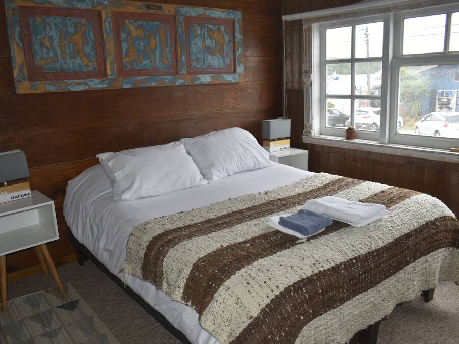 Une chambre avec un grand lit, deux tables de chevet, une fenêtre donnant sur des voitures garées et un mur orné d’œuvres d’art abstraites.