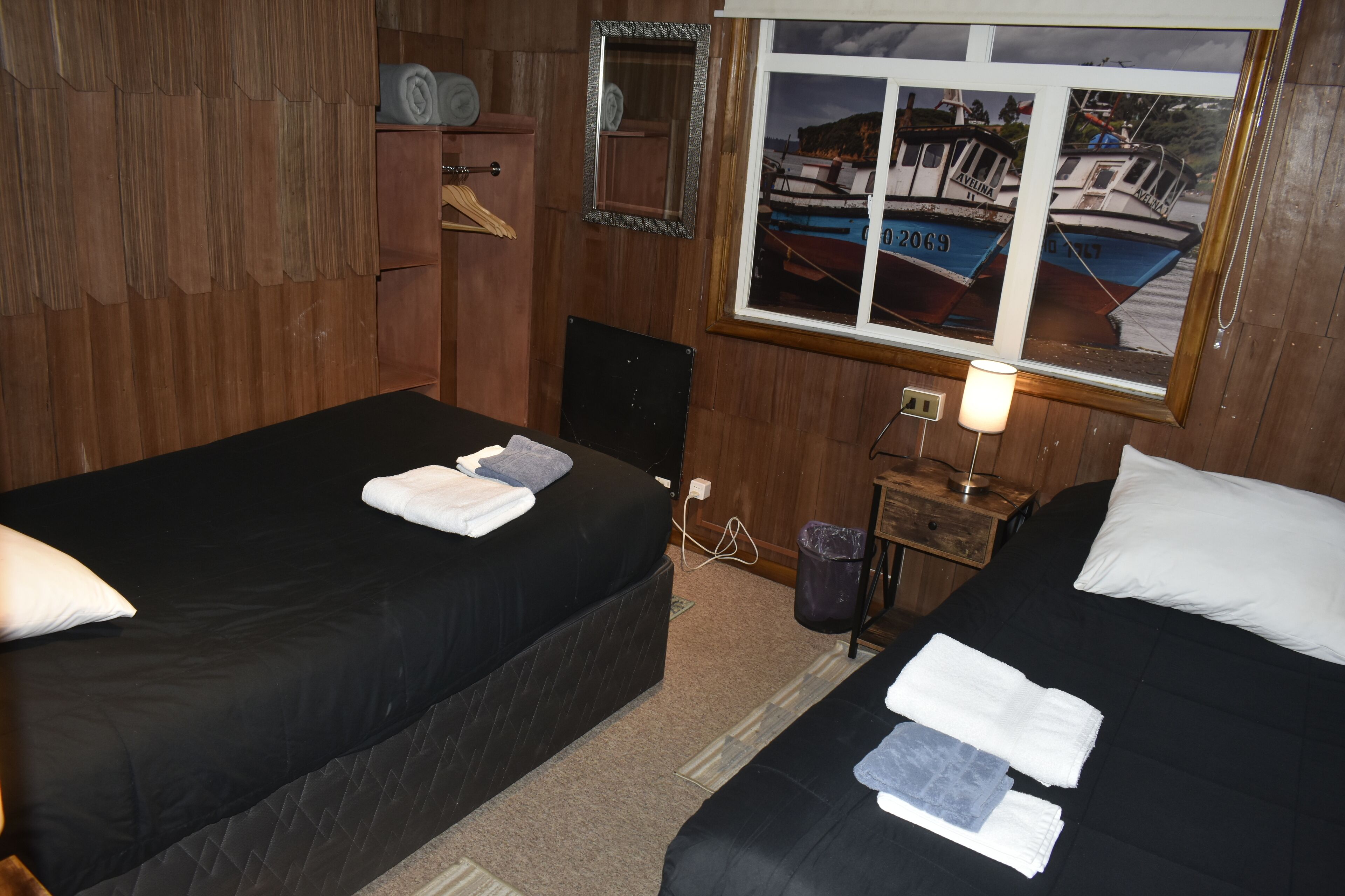 Una habitación con dos camas, paredes de madera, una ventana con vista a barcos, un espejo y una mesita con una lámpara.