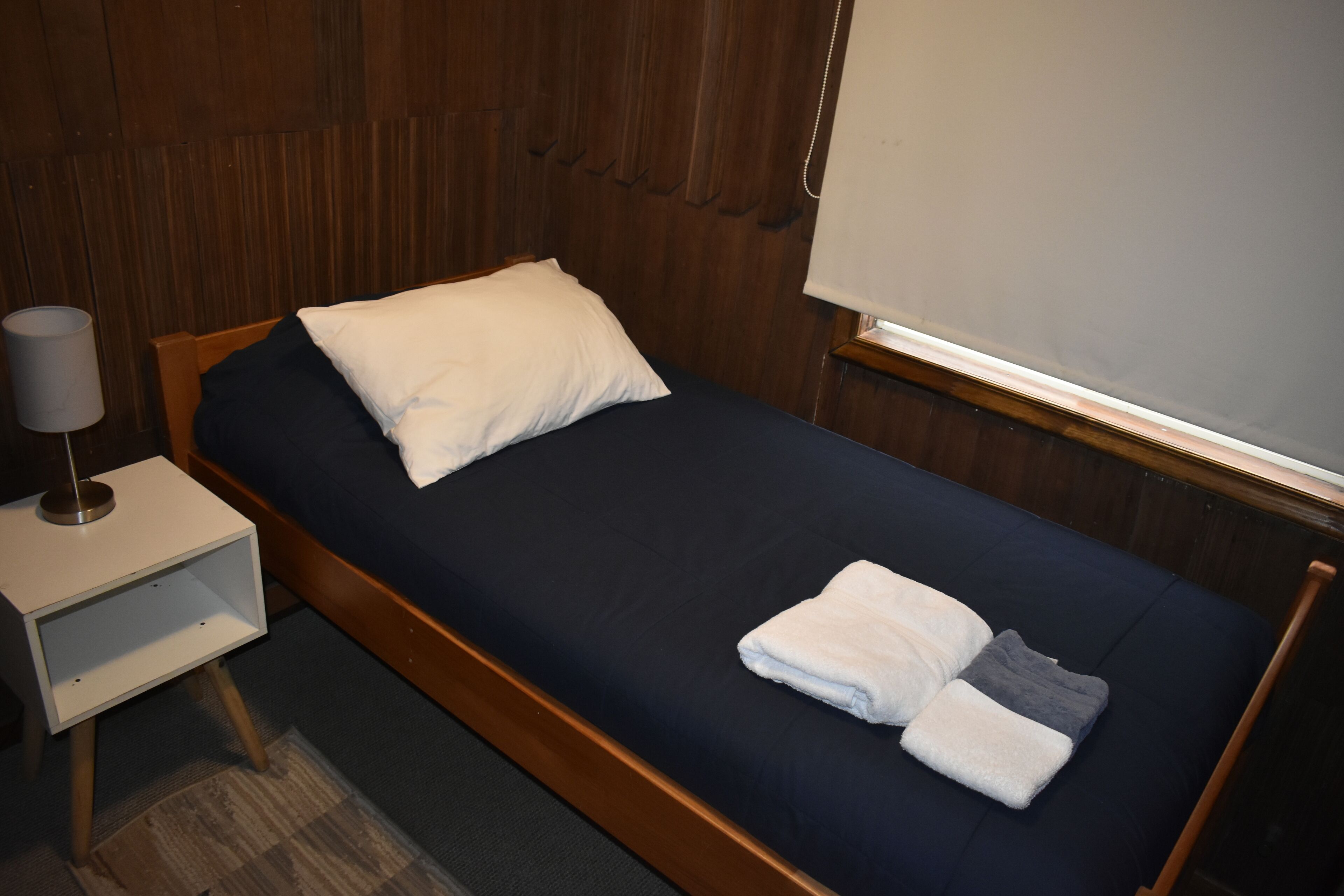 A single bed with a dark blue mattress, a white pillow, and a folded white towel. There is a bedside table with a lamp and a small shelf.