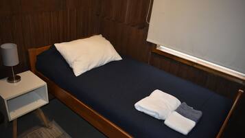 A single bed with a dark blue mattress, a white pillow, and a folded white towel. There is a bedside table with a lamp and a small shelf.