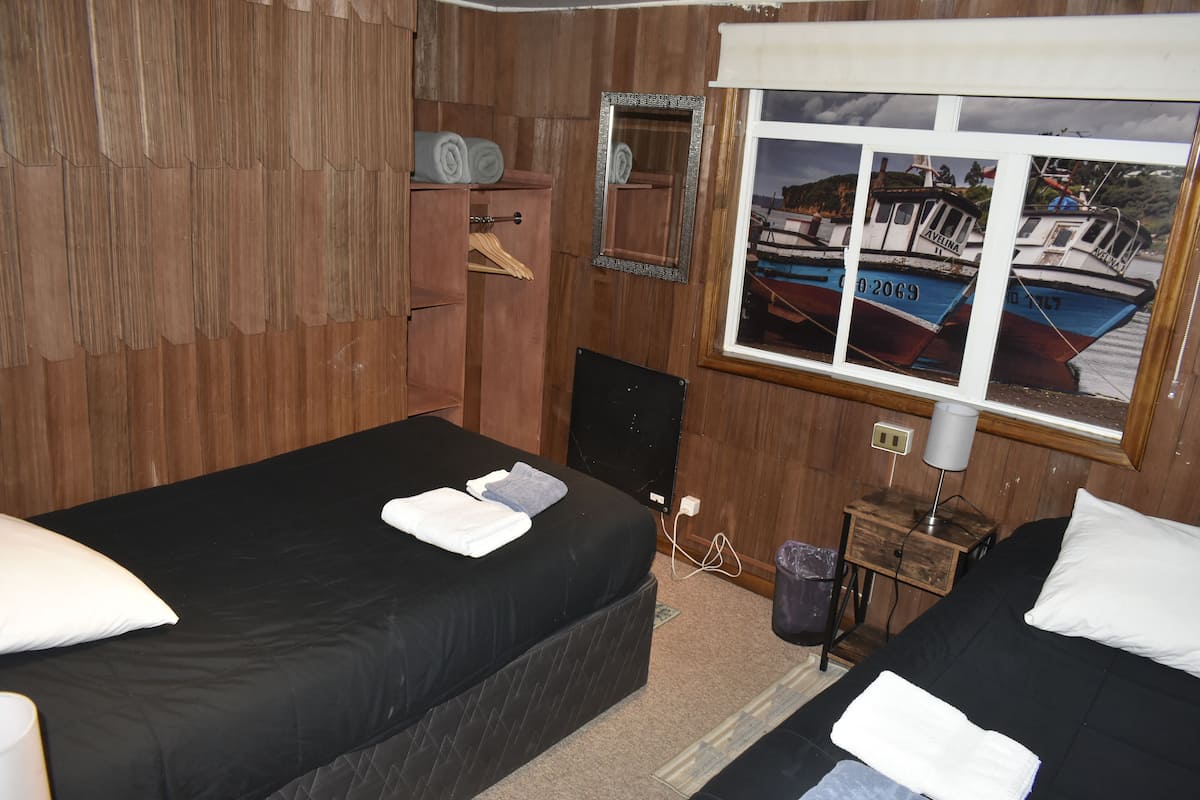 A room with two beds, wooden walls, a window with a view of boats, a mirror, and a small table with a lamp.
