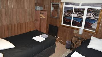 A room with two beds, wooden walls, a window with a view of boats, a mirror, and a small table with a lamp.
