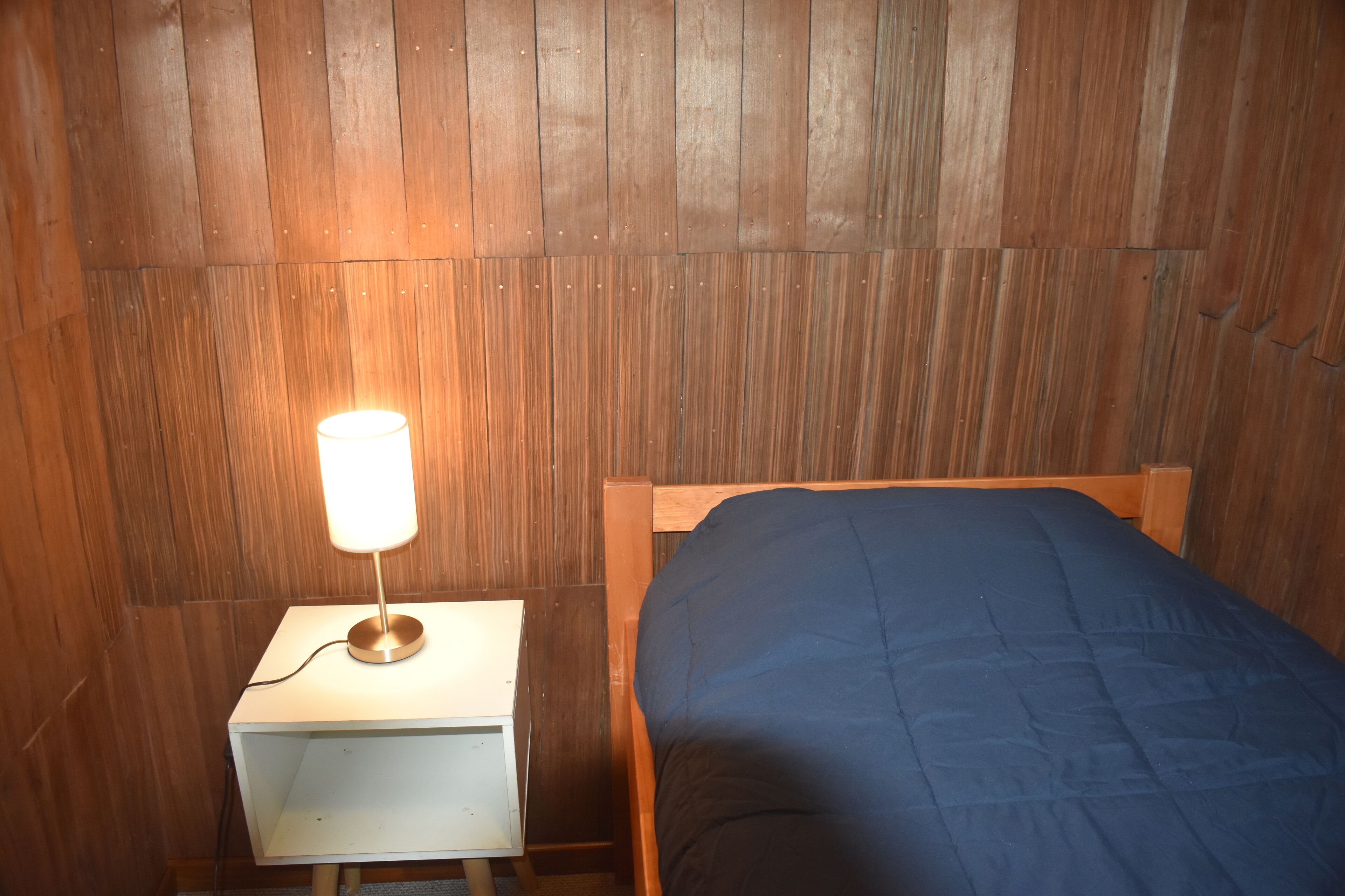 A wooden-paneled room with a bed and a bedside table with a lamp.