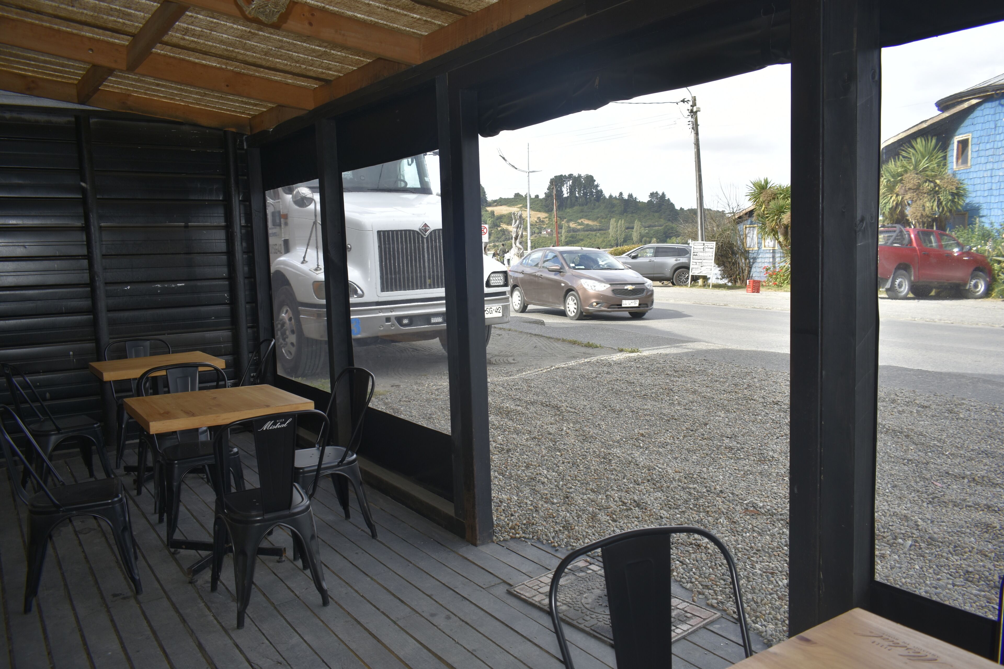 Een terras met tafels en stoelen, met uitzicht op een straat met geparkeerde auto's en een vrachtwagen, en een gebouw op de achtergrond.