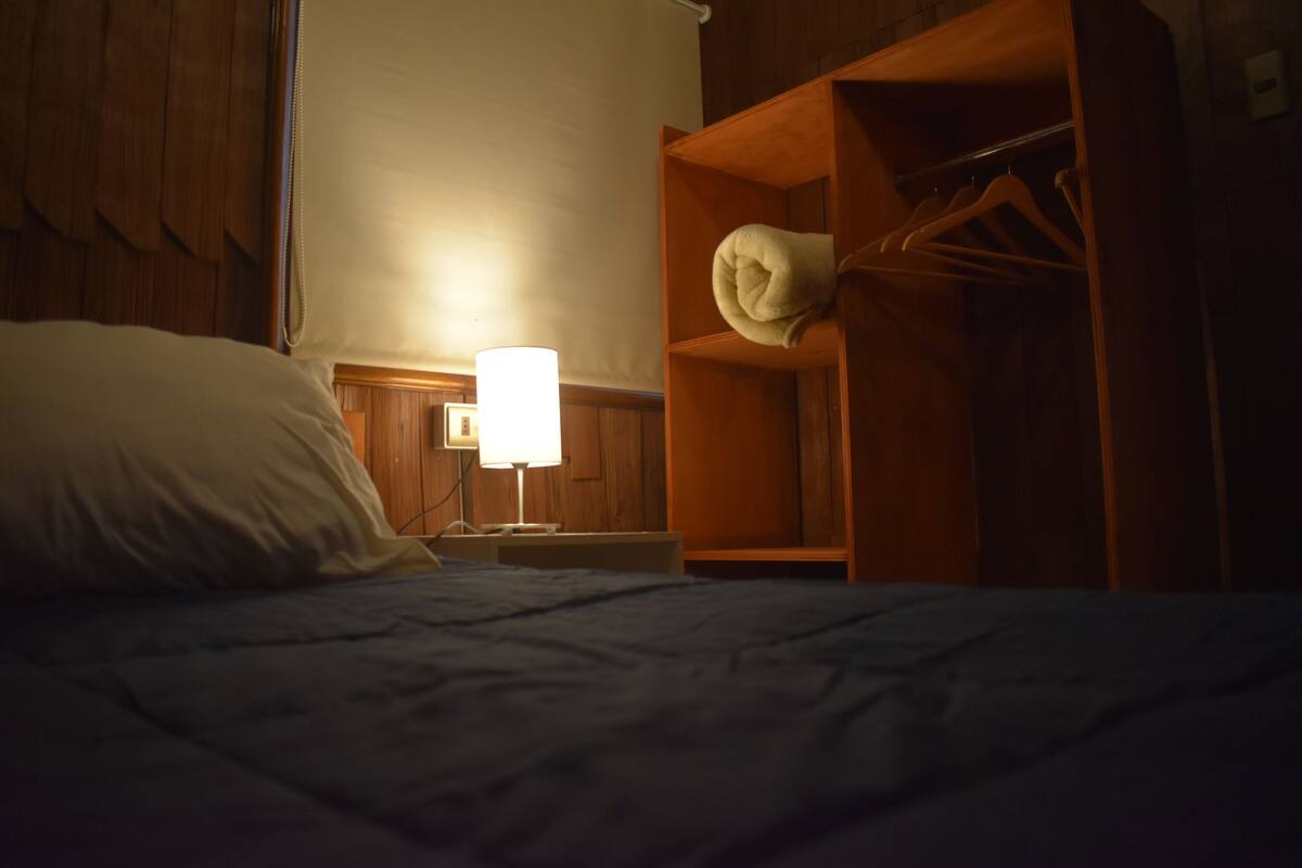 A dimly lit hotel room with a bed, a bedside lamp, a wooden wardrobe, and a wall-mounted shelf.