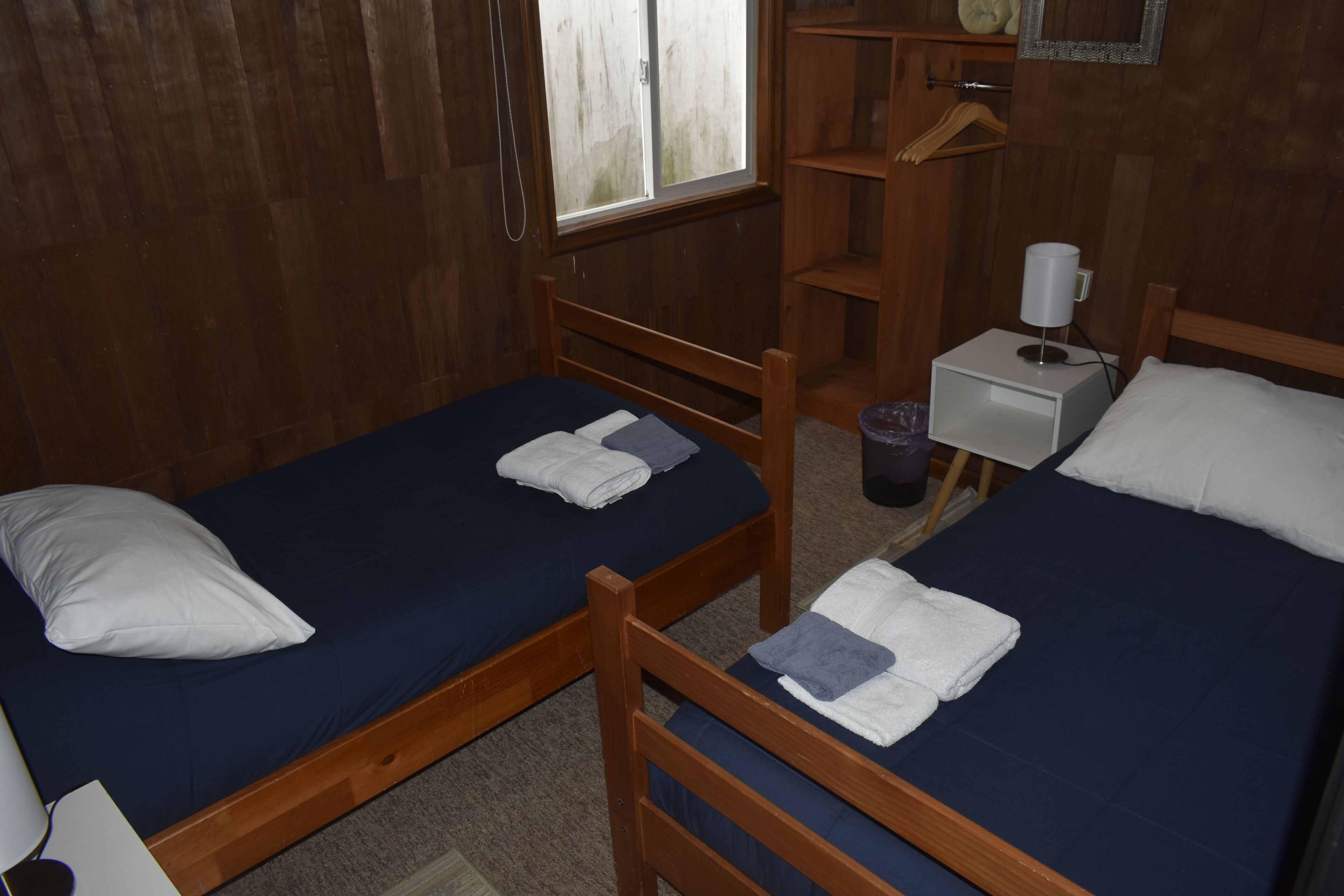 Una habitación con dos camas de madera, cada una con colchón azul y almohadas blancas. Hay una mesita de noche con una lámpara y un cesto de basura.