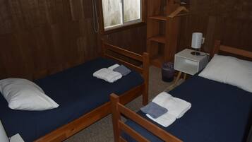 Una habitación con dos camas de madera, cada una con colchón azul y almohadas blancas. Hay una mesita de noche con una lámpara y un cesto de basura.