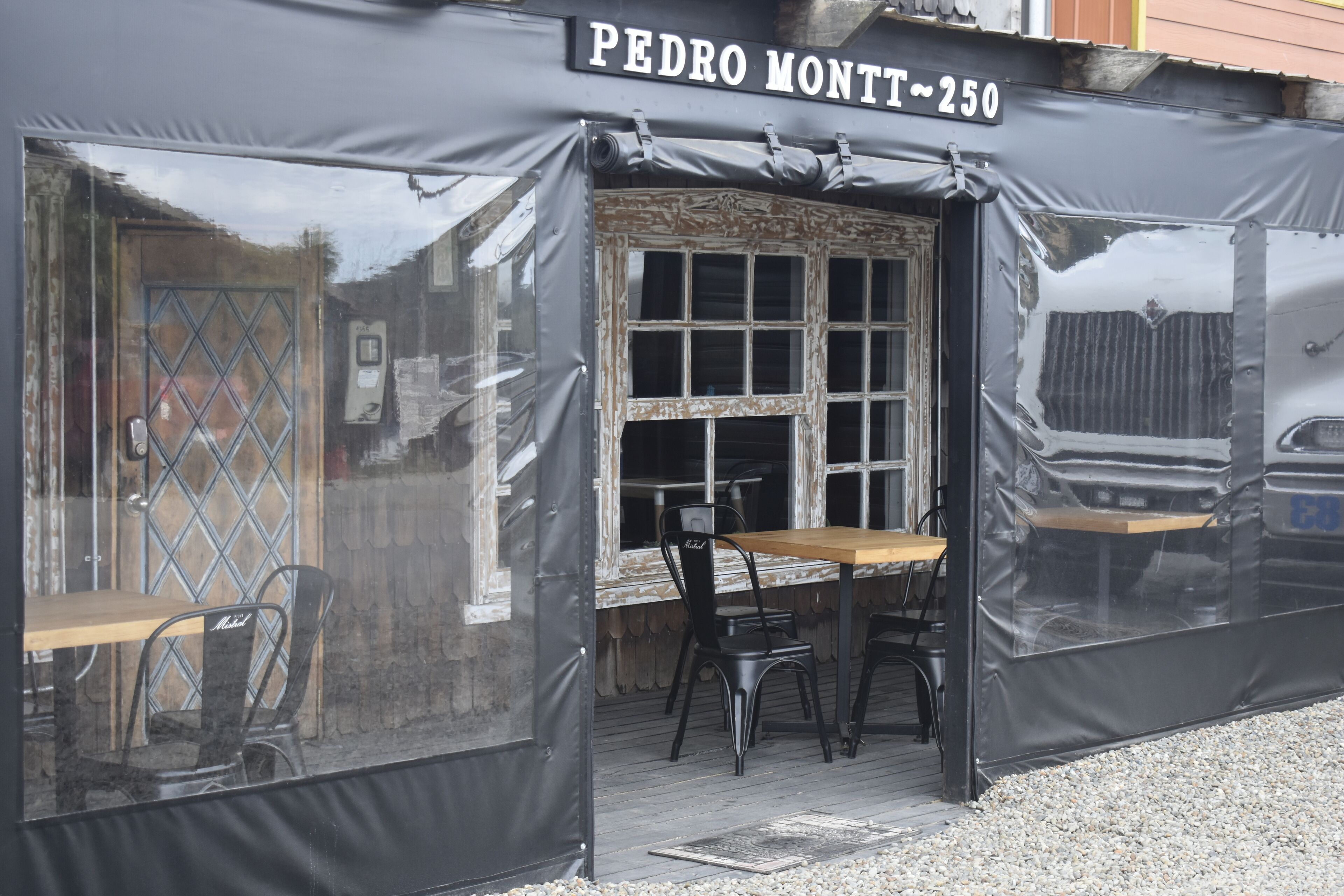 Un área de estar al aire libre cubierta con una mesa y sillas, una ventana con diseño de cuadrícula y un letrero que dice "PEDRO MONTT ~250".