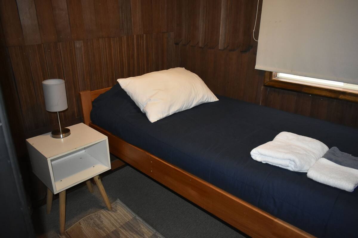 A single bed with a wooden frame, a white pillow, and a dark blue bedspread. There is a small bedside table with a lamp and a folded towel on the bed.