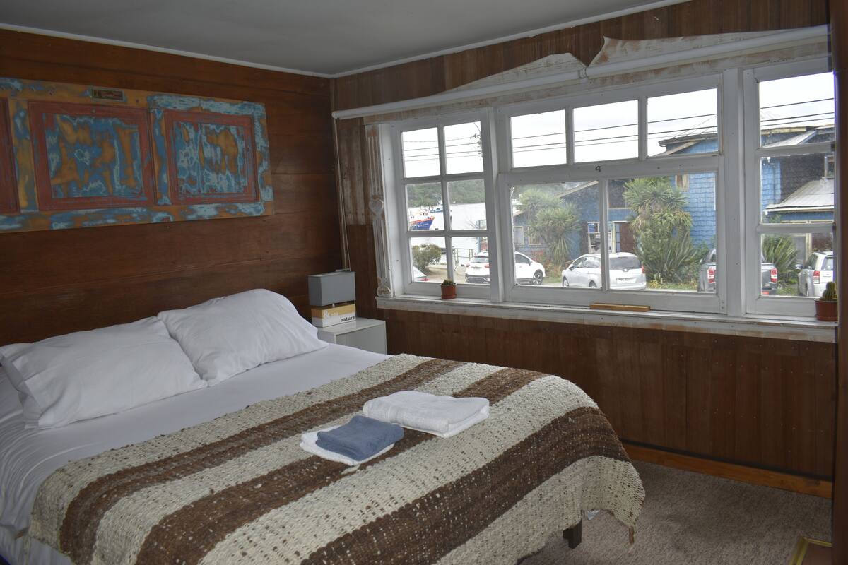 A bedroom with a large bed, a window with a view of parked cars and a building, and a wall with abstract art.