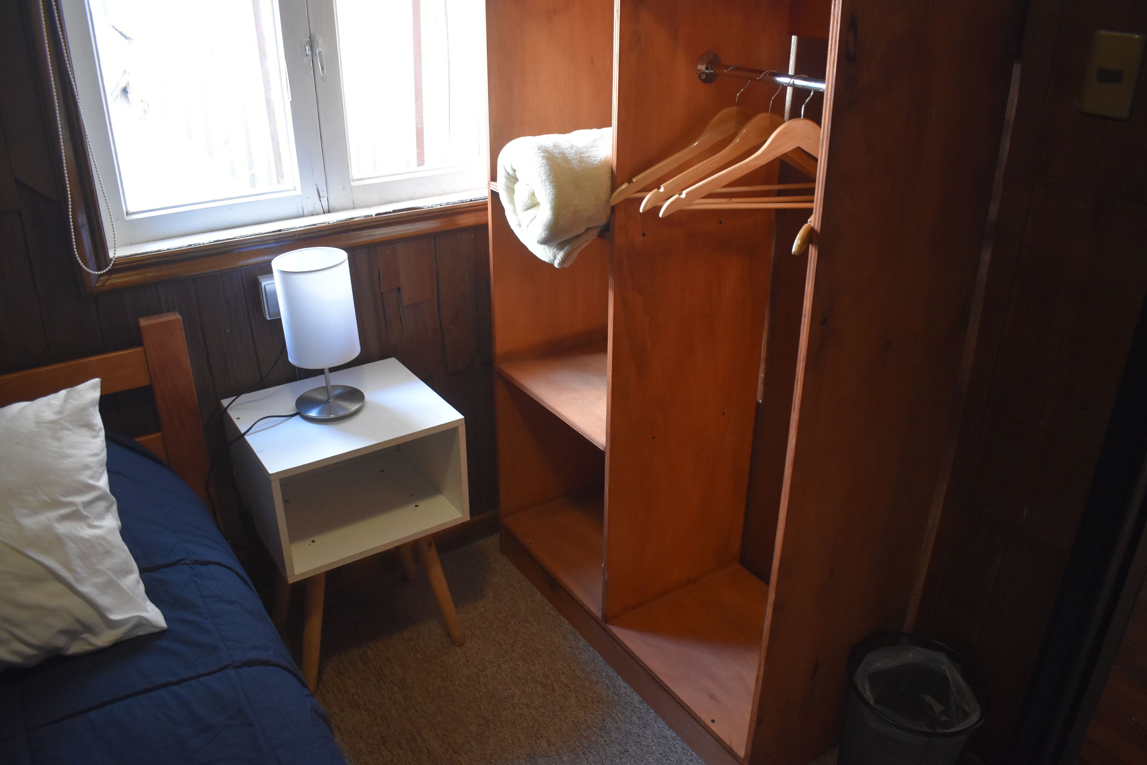 A bedroom with a bed, a bedside table with a lamp, a wooden wardrobe, and a window.