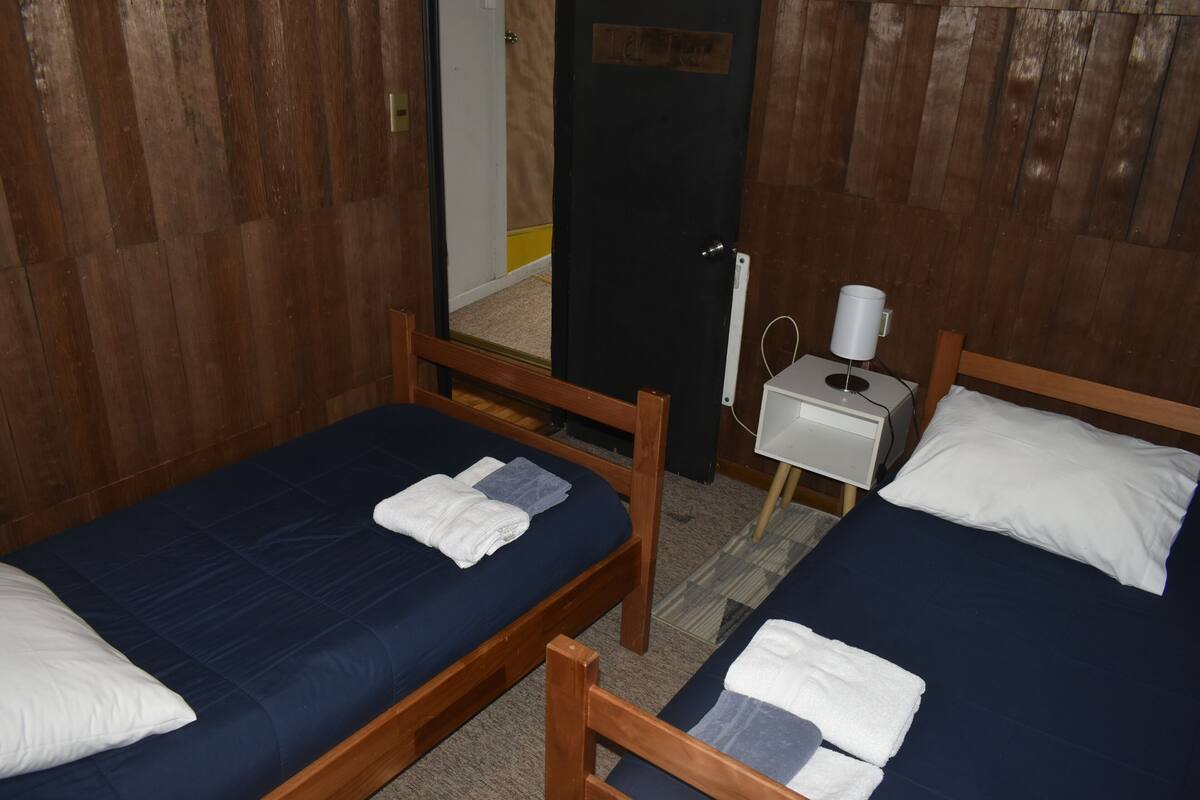 A room with two wooden beds, each with a blue mattress and white pillows. There is a small bedside table with a lamp and a power outlet.