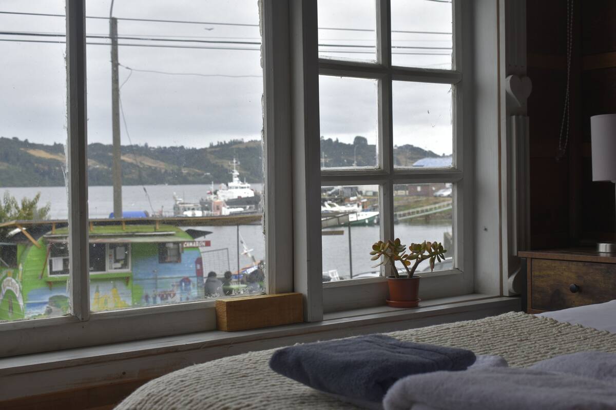 A room with a large window overlooking a harbor, a bed with a blue blanket, a wooden nightstand, and a potted plant.