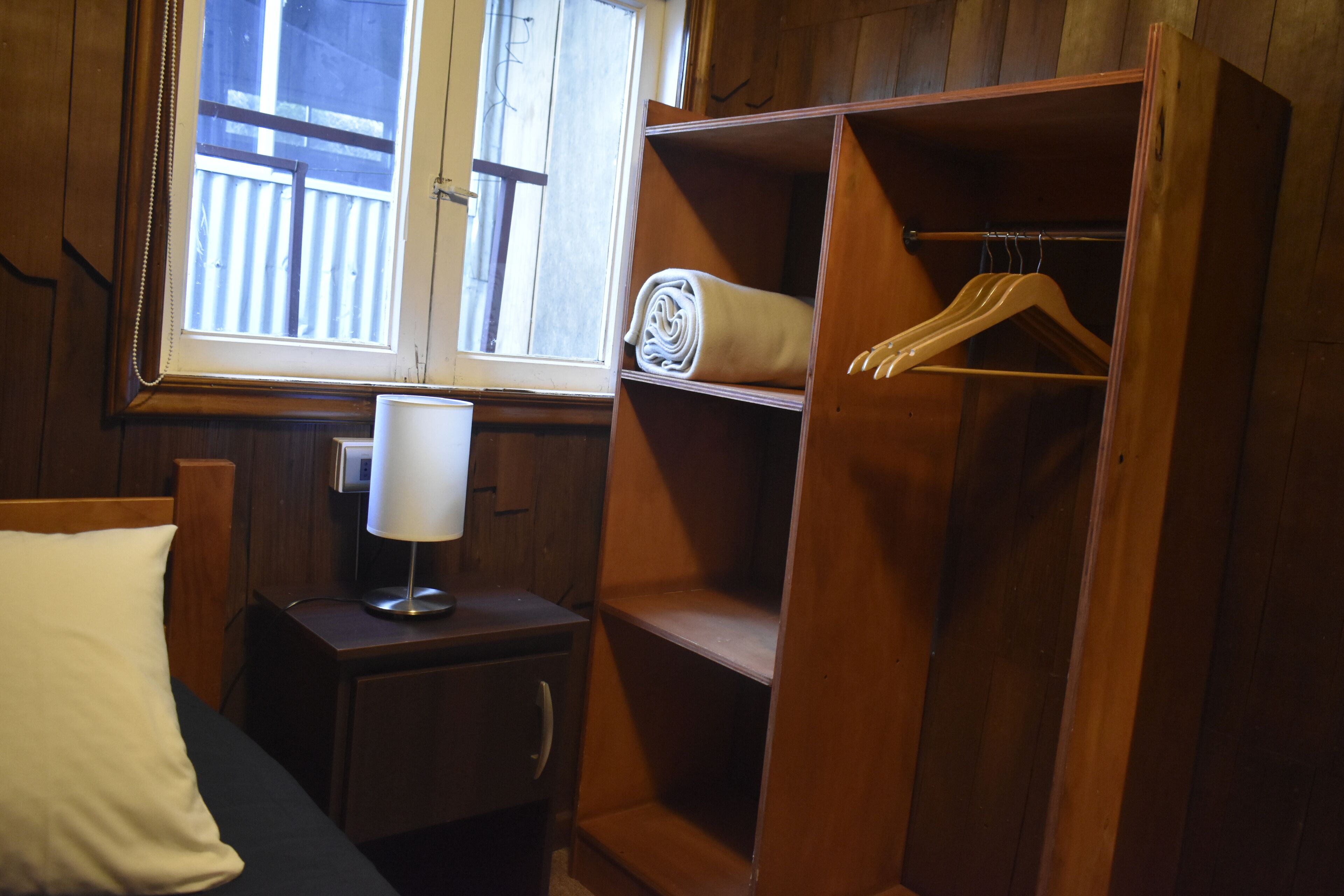 Una habitación de madera con una cama, una mesita de noche, una lámpara, una ventana y un armario de madera con perchas.
