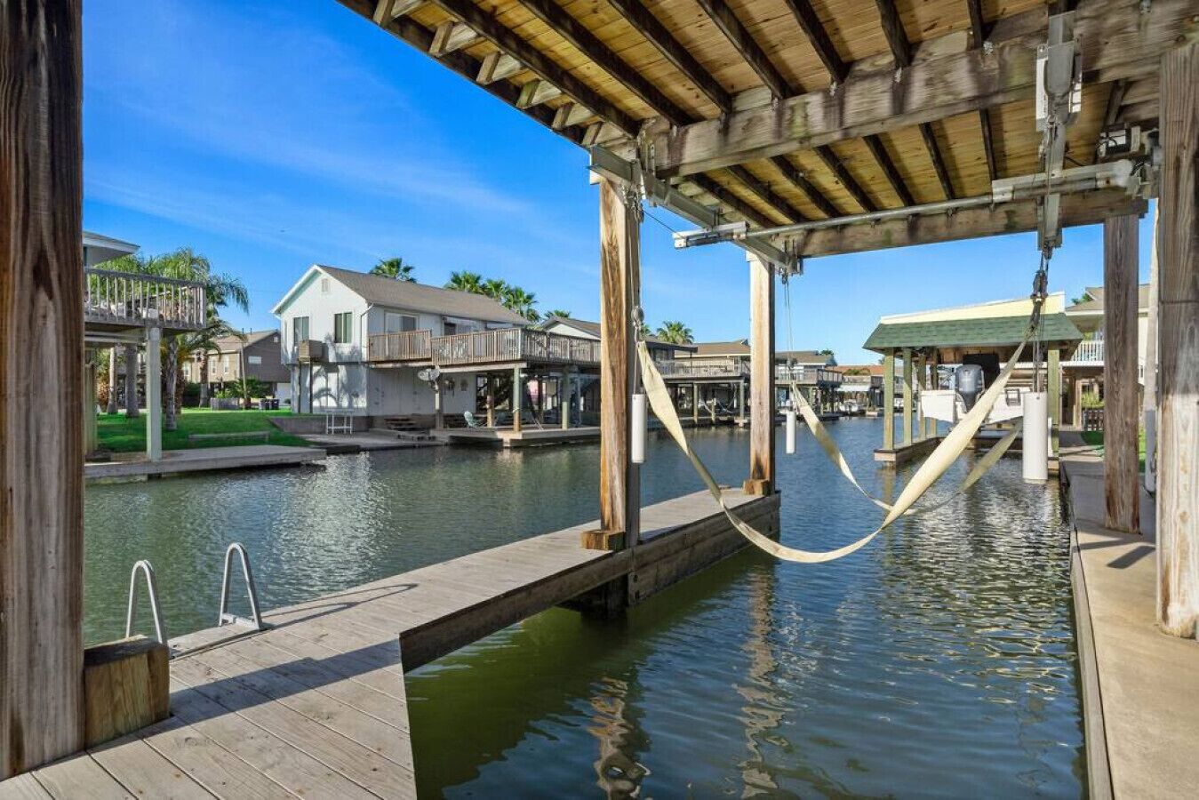 A wooden dock extending over water with a hammock, a house in the background, and a clear sky.