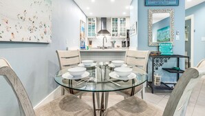 A dining area with a glass table surrounded by cushioned chairs, a kitchen in the background, and a decorative mirror on the wall. - Cassine- 3 Min to Beach, 2 BR, Pool, Bikes, Tennis & Beach (Santa Rosa Beach)