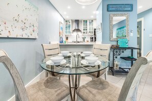 A dining area with a glass table surrounded by cushioned chairs, a kitchen in the background, and a decorative mirror on the wall. - Cassine- 3 Min to Beach, 2 BR, Pool, Bikes, Tennis & Beach (Santa Rosa Beach)