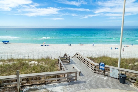 A wooden boardwalk leading to a beach with clear blue water, beach chairs, and umbrellas.