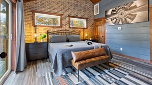 Une chambre avec un mur en briques, un lit avec une banquette, une fenêtre et des œuvres d’art accrochées au mur.