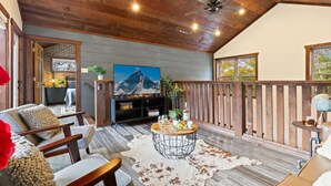 Un salon chaleureux avec un plafond en bois, un téléviseur fixé au mur, une table à manger avec des chaises et une vue sur l’extérieur grâce à de grandes fenêtres.